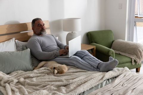 Mature man working on laptop in cozy bed with sleeping golden retriever puppy