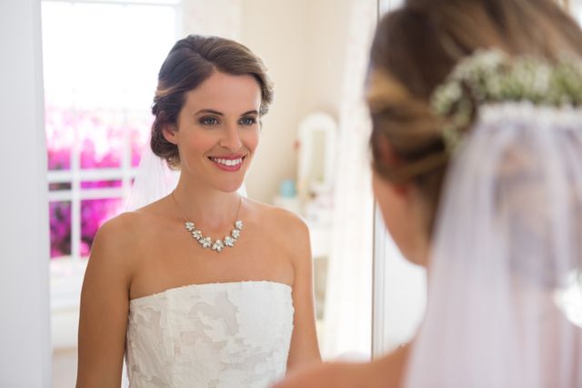 This image captures a bride smiling at her reflection in a mirror, showcasing her wedding dress and accessories. Ideal for use in wedding planning websites, bridal magazines, and advertisements for bridal boutiques. It conveys themes of joy, beauty, and anticipation.