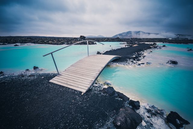 Wooden bridge extends over strikingly blue thermal waters with volcanic rocks around. Mist rises in the background, creating a soothing ambiance amid vast rocky terrain. Ideal for travel blogs, nature magazines, and promotional materials for vacation and tour packages focusing on Iceland. Conveys tranquility and unique outdoor adventure.