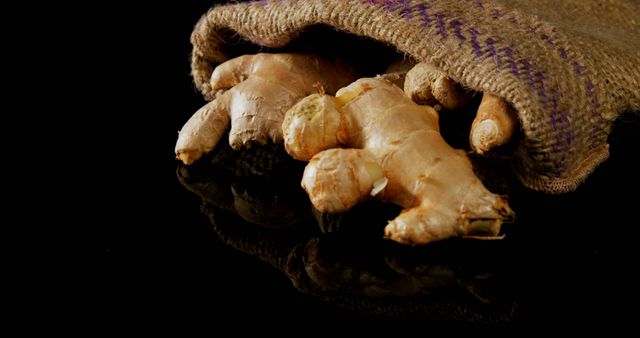 A fresh ginger root spills out of a rustic burlap sack onto a reflective black surface, with copy space. Ginger is widely used for its flavor and medicinal properties in various cuisines and herbal remedies.