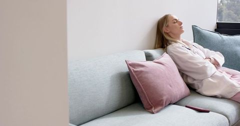 Young woman relaxing on pastel sofa in loungewear with pink cushion and remote, calm modern interior