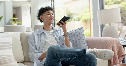 Asian Teen Male Relaxing on Sofa Speaking into Smartphone in Bright Modern Living Room