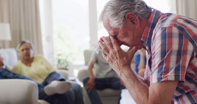Senior man looking distressed holding a tissue in a support group setting. Others sit around, offering community support. Useful for depicting mental health scenarios, elderly wellness, and group therapy sessions.