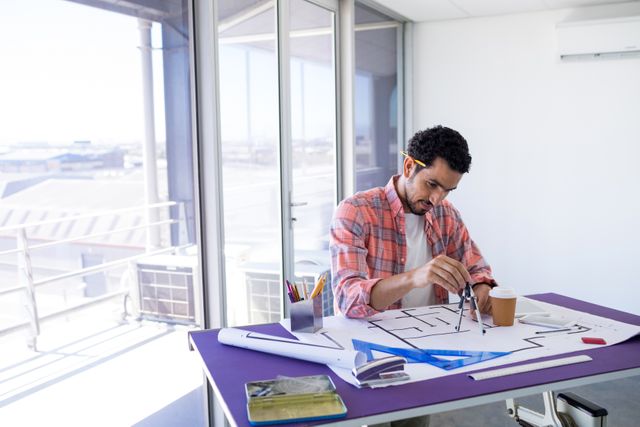 Male architect working on a blueprint over a drafting table in a modern office. Ideal for use in articles or advertisements related to architecture, engineering, construction, design projects, and professional work environments.