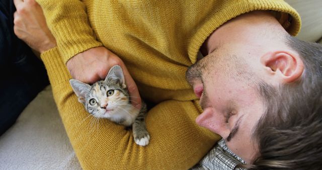 Man lying on couch happily cuddling with a small kitten while wearing a yellow sweater. Great for use in articles or ads related to pet adoption, animal companionship, home comfort, mental well-being, and the bond between humans and their pets.