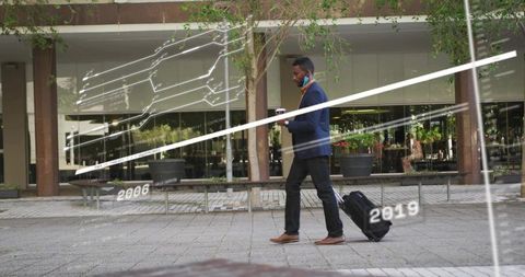 Business commuter walking with rolling suitcase and coffee through urban plaza reflections