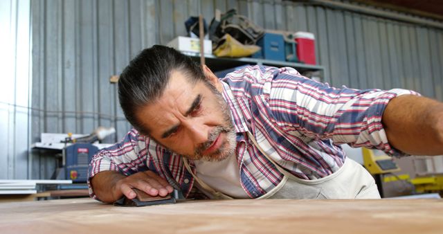 Mature male carpenter sanding wooden board in a workshop, wearing apron and checkered shirt. Ideal for use in articles or advertisements related to woodworking, craftsmanship, DIY projects, professional skills or wood industry. The focused expression and detailed hands-on work illustrate dedication and expertise.