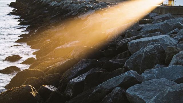 Golden sunbeam cutting across rocky breakwater with white beacon at shoreline