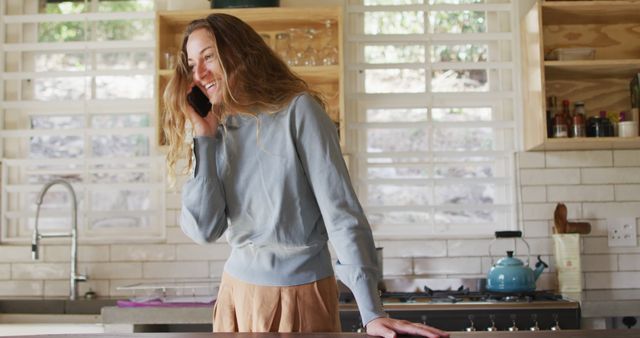 Happy caucasian woman standing in sunny cottage kitchen talking on smartphone and smiling. simple living in an off the grid rural home.