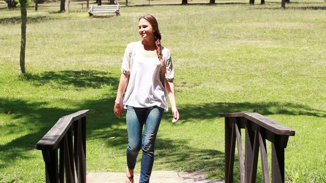 Woman walking across small bridge in open park area on a sunny day. She is wearing casual clothing including jeans and a t-shirt. Surrounding is a green grassy field with scattering of trees. Perfect video for themes related to nature walks, outdoor activities, leisure time, relaxation, and healthy living.