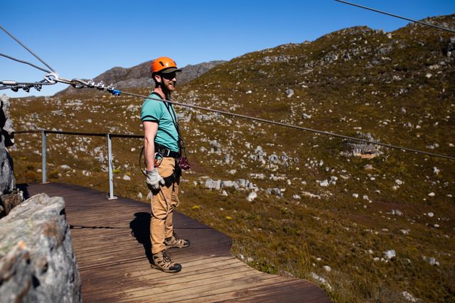 Caucasian man standing on wooden decking, wearing zip lining equipment and helmet, admiring mountainous landscape on a sunny day. Perfect for promoting adventure tourism, outdoor activities, and vacation destinations.