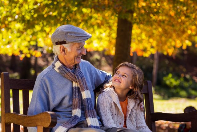 Grandfather and granddaughter enjoying a moment together on a park bench during autumn. Ideal for use in family-oriented advertisements, articles about generational bonding, and promotions for outdoor activities or senior living.