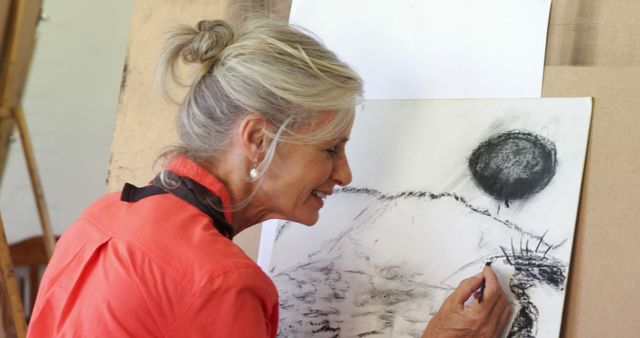 An elderly woman is seen drawing a landscape sketch in an art studio, showcasing her artistic talent and passion for creativity. This image can be used for themes related to senior activities, hobbies, creativity, and aging gracefully. Ideal for articles on art therapy, retirement activities, or promotions for art classes aimed at seniors.