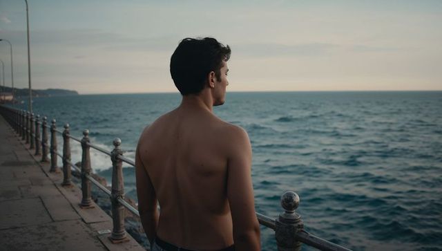 Shirtless man standing on coastal promenade looking toward ocean at dusk, solitude seascape