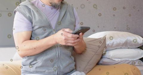Mature asian man checking smartphone on bed wearing grey hoodie with pillows and soft bokeh