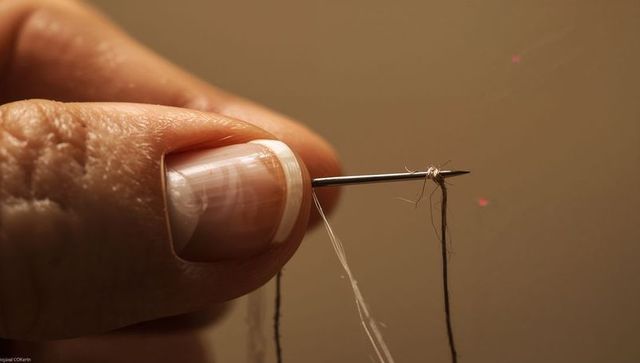 Macro fingertip holding threaded needle with knotted dark thread and French manicure