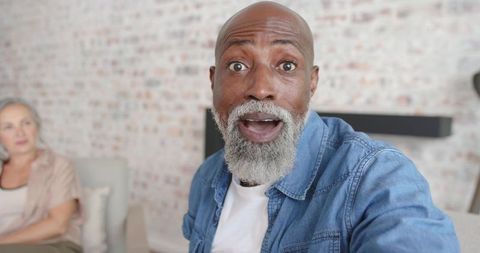 Senior man reacting with astonished expression in cozy living room with partner on sofa