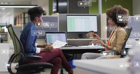 Two women collaborating over spreadsheets in open office with social media data overlays
