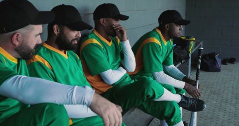 Pensive multicultural baseball players sitting in dugout wearing green and yellow uniforms