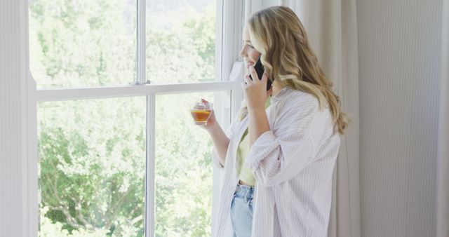 Woman standing by bright window, holding hot beverage and talking on phone. Ideal for depicting everyday lifestyle, morning routines, work-from-home scenarios, or relaxation scenes.