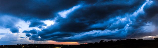 This panoramic capture of a sunset with dark storm clouds presents an atmospheric and dramatic outdoor scene. The deep blue and black clouds contrast vividly with the warm orange horizon, indicating an impending storm during twilight. This image is ideal for use in content related to weather forecasting, nature, climate change, storm warnings, backgrounds for presentations, or decorative art for those appreciating the power and beauty of nature.