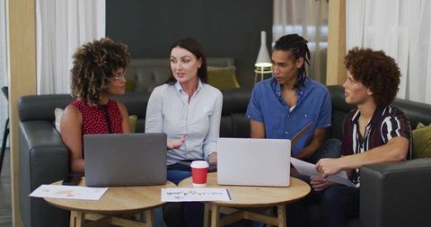 Woman leading diverse team in informal lounge meeting with laptops, charts, collaboration