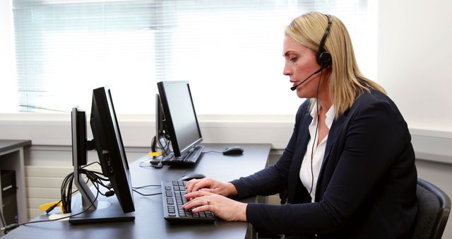 A Caucasian middle-aged woman works diligently at her desk in a customer service environment, with copy space. She is equipped with a headset, suggesting she is engaged in handling calls or providing remote assistance.