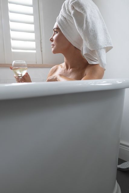 This image depicts a woman enjoying a moment of relaxation in a bathtub, holding a glass of champagne. She has a towel wrapped around her head, suggesting a spa-like experience. This image can be used for promoting self-care, luxury spa services, wellness products, or relaxation techniques. It is ideal for websites, blogs, or advertisements focused on health, beauty, and lifestyle.