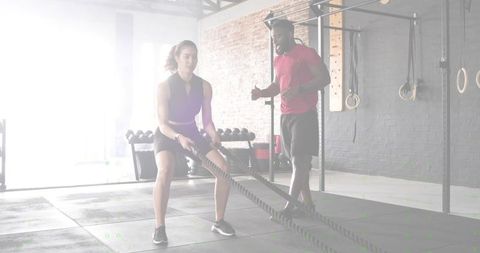 Female athlete performing battle rope squat with trainer coaching in brick wall functional gym
