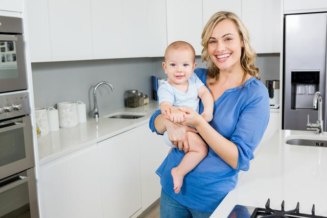 Mother in a modern, white kitchen holding her infant. She is smiling, depicting warmth and happiness. Ideal for use in advertising and articles about parenting, family life, home environment, and domestic activities.