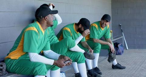 Multiracial baseball team relaxing in dugout laughing and chatting in green uniforms