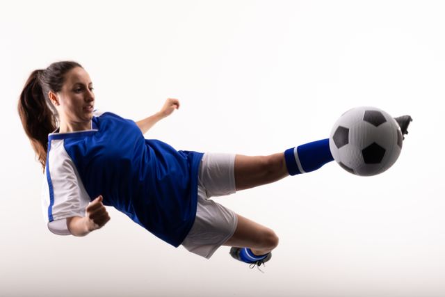 Young female soccer player in blue and white sportswear kicking a soccer ball mid-air against a white background. Ideal for use in sports-related content, advertisements for athletic gear, fitness promotions, and articles about women's sports.