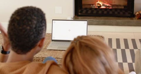 Diverse couple sitting on sofa watching laptop by fireplace, cozy home relaxation