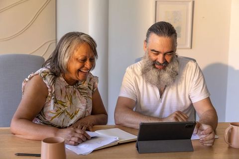 Senior couple laughing and reviewing notes while using tablet at home