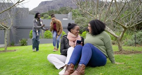 Black friends laughing on lawn while two women sharing phone during casual outdoor hangout