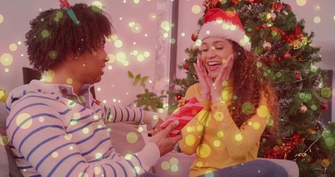 Couple exchanging christmas gift on couch by decorated tree, joyful holiday moment