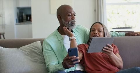 Mature african american couple sharing smartphone and tablet while relaxing on sofa
