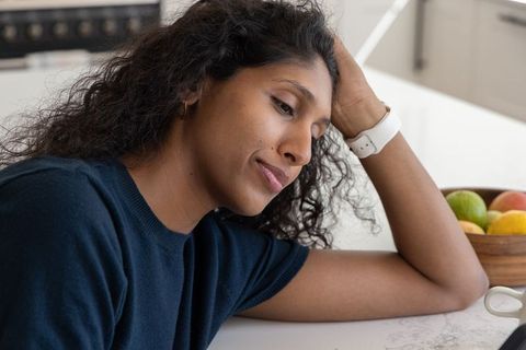 Contemplative woman leaning on kitchen counter wearing smartwatch near fruit bowl