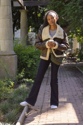 Sunlit garden style african woman wearing shearling jacket holding coffee and crossbody bag