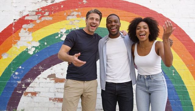 Laughing friends standing in front of rainbow mural wearing jeans, urban togetherness