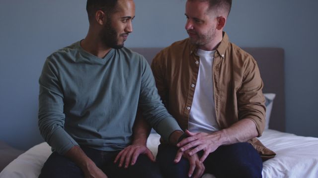 Two men, one with a darker skin tone and the other with a lighter skin tone, are sitting on a bed and showing affection towards each other. The video depicts a loving and intimate moment between a same-sex couple. This video can be used in campaigns promoting LGBTQ inclusiveness, representations of interracial relationships, and home life. It is suitable for articles, blogs, advertisements, and social media posts celebrating love, diversity, and togetherness.