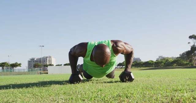 Fit african american man exercising outdoors doing press ups holding dumbbells. cross training for fitness at a sports field.