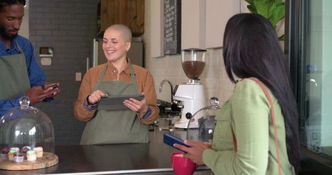 Smiling barista using tablet serving indian customer with phone at coffee counter