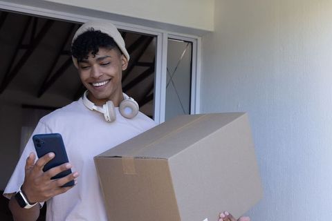 African american man in 20s carrying parcel while checking smartphone at doorway