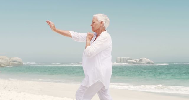 Senior woman practicing Tai Chi on beach with ocean background. Ideal for use in materials related to healthy aging, fitness routines, relaxation techniques, wellness programs for seniors, and stress reduction. Represents serenity, balance, and physical exercise for elderly individuals.