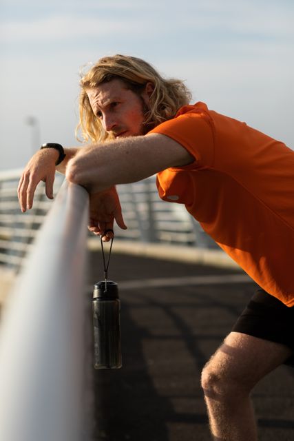 Side view of a fit Caucasian man with long blonde hair wearing sportswear exercising outdoors in the city on a sunny day with blue sky, standing and resting on a footbridge, holding water bottle in hand.