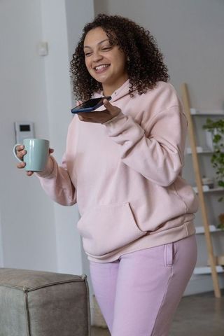 Smiling african american woman in pastel hoodie holding mug and phone, home lifestyle