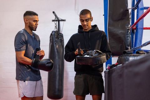 Boxing partners adjusting gloves and inspecting focus mitts beside heavy bag in gym ring