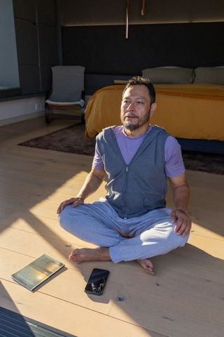 Asian man sitting cross-legged reaching for smartphone in minimalist bedroom sunlight