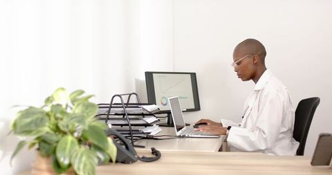 Medical professional typing on laptop at minimalist bright office desk with charts